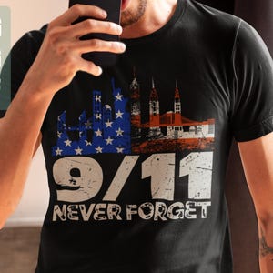 May include: Black t-shirt featuring a distressed graphic design with the text "9/11 NEVER FORGET." The design incorporates the American flag, the Twin Towers, and a bridge. The shirt is a crew neck.