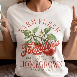 May include: White t-shirt with a vintage-style graphic design featuring a tomato vine with red tomatoes and the text "FARM FRESH Tomatoes SINCE 100% ORGANIC 1976 HOMEGROWN PICK YOUR OWN".