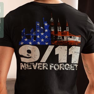 May include: Black t-shirt featuring a distressed graphic commemorating 9/11. The design includes a skyline with the Statue of Liberty and other buildings, with the American flag colors. The text "9/11 NEVER FORGET" is prominently displayed.