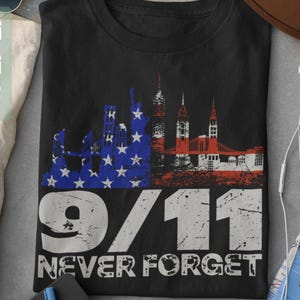 May include: Black t-shirt with a distressed graphic of a skyline and American flag design. The text "9/11 Never Forget" is printed below the graphic in white. The shirt is a tribute to the events of September 11th.