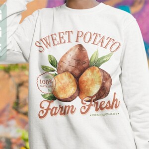 May include: White sweatshirt with a brown and orange graphic design of sweet potatoes with leaves and the text "Sweet Potato Farm Fresh - Premium Quality" and a 100% natural label.
