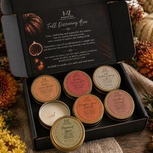 May include: A black cardboard box containing six 2 oz soy wax candles in round gold tins. The box lid features the text Fall Discovery Box and Modern Zen Candle Company. The candles are labelled Autumn Creme Brulee, Bourbon Evening Glow, Fall Frosting Breeze, Pumpkin Spice Delight, Woodland Harvest, and Pearwood Spice.