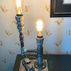 May include: A silver model car sits on a wooden base with a metal top. The base has a switch and two exposed wires. Two Edison light bulbs are attached to a pipe lamp fixture.