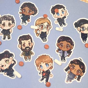 Puede incluir: Una colección de nueve pegatinas troqueladas con personajes de estilo chibi en varios atuendos, incluyendo uniformes médicos y ropa casual, esparcidas sobre una superficie azul claro junto a varias pastillas pequeñas.