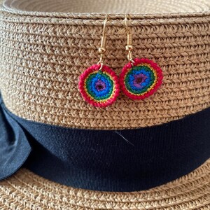 May include: A pair of crocheted earrings with a rainbow and blue color scheme. The earrings are hanging from a straw hat.