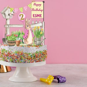 May include: A white cake with colorful sprinkles decorated with a bunny rabbit, a birdhouse, a fence, flowers, and a sign that says "Happy Birthday ESME". The cake is on a white cake stand and there are party favors in the foreground.