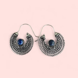 Könnte beinhalten: Ein Paar silberfarbene Creolen mit aufwendigen Filigranarbeiten. Jeder Ohrring hat eine halbmondförmige Form mit einem zentralen, runden, dunkelblauen Stein. Die Ohrringe haben einen Hakenverschluss, vor hellrosa Hintergrund.