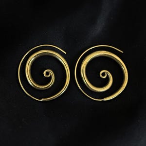 Può includere: Un paio di orecchini in metallo color oro a forma di spirali aperte. Ogni orecchino presenta un filo continuo che si avvolge verso l'interno da un bordo esterno più ampio fino a un punto centrale stretto, creando una silhouette circolare.