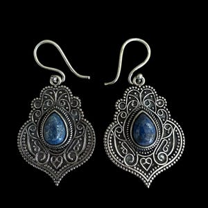 Könnte beinhalten: Ein Paar silberfarbene Ohrringe mit einem tropfenförmigen blauen Edelstein in der Mitte. Die Ohrringe haben ein verziertes Filigran-Design mit Hakenverschluss. Der Hintergrund ist einfarbig schwarz.