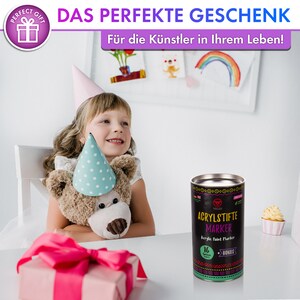 Puede incluir: Una lata negra y verde con el texto "VELLES ACRYLSTIFTE MARKER Acrylic Paint Marker 16 Farben + BONUS" en ella. La lata est&aacute; sobre una mesa blanca con una caja de regalo rosa y un oso de peluche que lleva un gorro de fiesta azul y blanco.