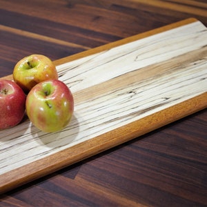 May include: A long, rectangular wooden serving board with a natural wood grain pattern. The board has a light brown border and a handle on one end. Three red and green apples are placed on the board.
