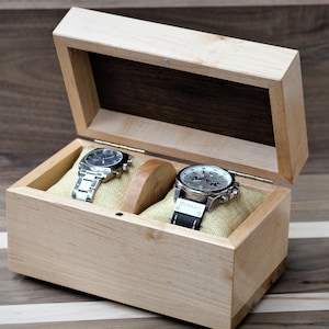 May include: A wooden watch box with a hinged lid, open to reveal two watches on cushions. The box is made of light wood with a dark wood interior.