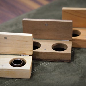 May include: Three wooden ring boxes with hinged lids. The boxes are made from different types of wood, and each has two compartments for rings. The boxes are arranged in a row, with the largest box at the back and the smallest box at the front.