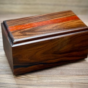 May include: A rectangular wooden box with a lid, featuring a rich, dark brown grain. A vibrant orange-red stripe accents the top. The box has a classic, elegant design, perfect for storing small items.