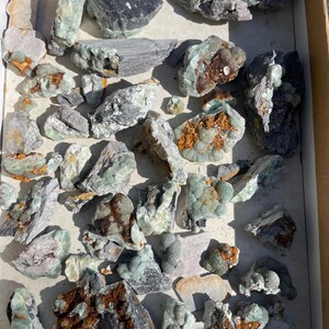 May include: A collection of raw, uncut gemstones in a cardboard box. The stones vary in size and shape, displaying a range of colors including shades of green, gray, brown, and black. Some stones have a crystalline texture, while others appear more rough and natural.