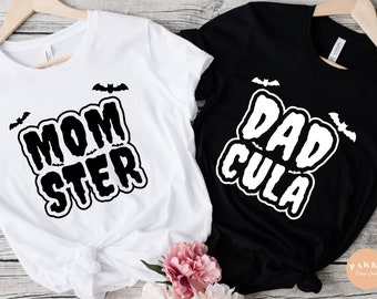 May include: Two black and white t-shirts with a Halloween theme. The white t-shirt says "Momster" with bats above and below the text. The black t-shirt says "Dadcula" with bats above and below the text.