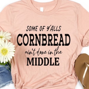 Puede incluir: Una camiseta rosa con el texto "Some of y'alls cornbread ain't done in the middle" impreso en negro.
