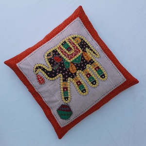 May include: A decorative cushion with an orange border and a white background features an embroidered elephant design with colourful patchwork fabric. The elephant is holding a ball in its trunk.