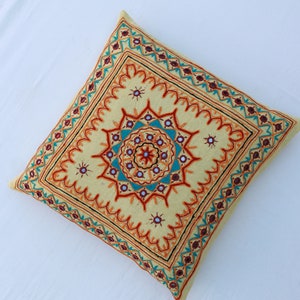 May include: A square yellow throw pillow with intricate embroidery in shades of orange, red, blue, and green. The embroidery features a central starburst design with smaller stars and geometric patterns.
