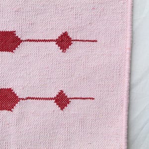 Puede incluir: Textil tejido rosa con un patr&oacute;n repetitivo de formas geom&eacute;tricas rojas. El dise&ntilde;o incluye elementos rectangulares y en forma de diamante. La tela tiene una apariencia texturizada, lo que sugiere una calidad artesanal o hecha a mano.