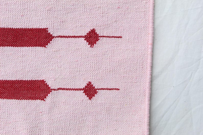 Puede incluir: Textil tejido rosa con un patr&oacute;n repetitivo de formas geom&eacute;tricas rojas. El dise&ntilde;o incluye elementos rectangulares y en forma de diamante. La tela tiene una apariencia texturizada, lo que sugiere una calidad artesanal o hecha a mano.