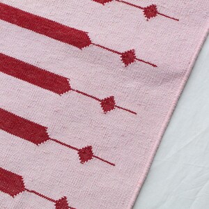 Puede incluir: Primer plano de un textil rosa y rojo estampado. El dise&ntilde;o presenta amplias rayas rojas con detalles en forma de diamante conectados por l&iacute;neas finas. El textil tiene una textura tejida, con los elementos rojos contrastando con el fondo rosa.
