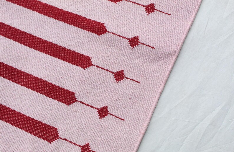 Puede incluir: Primer plano de un textil rosa y rojo estampado. El dise&ntilde;o presenta amplias rayas rojas con detalles en forma de diamante conectados por l&iacute;neas finas. El textil tiene una textura tejida, con los elementos rojos contrastando con el fondo rosa.