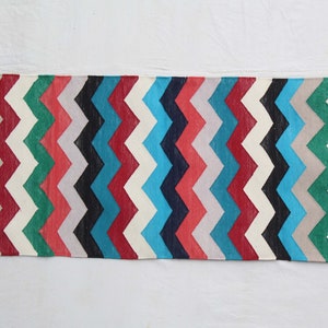 Op de afbeelding: Een rechthoekig tapijt met een zigzagpatroon in verschillende kleuren. Het tapijt heeft strepen in rood, wit, zwart, grijs, groen en blauw. Het is gemaakt van geweven stof en wordt waarschijnlijk gebruikt als decoratieve vloerbedekking.
