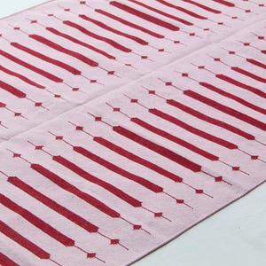Puede incluir: Una alfombra rosa con un patr&oacute;n repetitivo de rect&aacute;ngulos alargados y rombos rojos. El dise&ntilde;o es sim&eacute;trico, con los elementos rojos espaciados uniformemente sobre el fondo rosa. La alfombra es probablemente de tela.