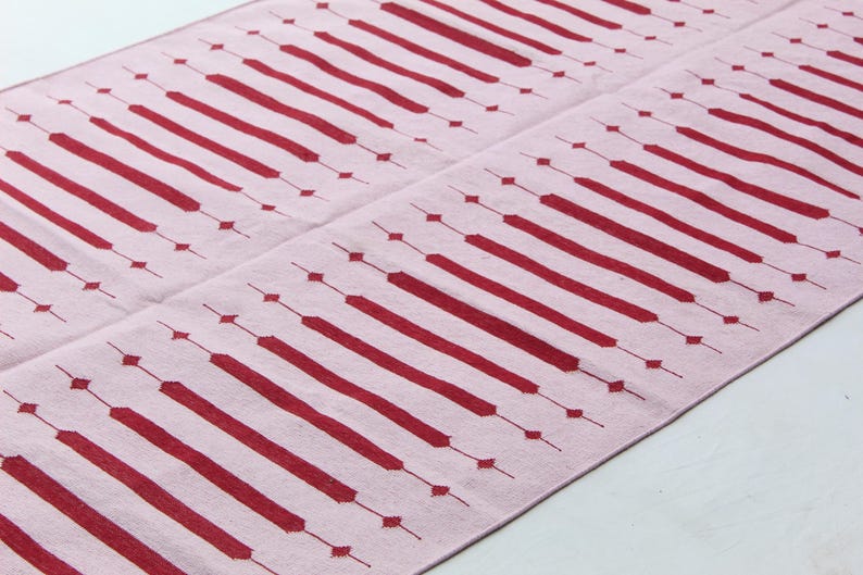 Puede incluir: Una alfombra rosa con un patr&oacute;n repetitivo de rect&aacute;ngulos alargados y rombos rojos. El dise&ntilde;o es sim&eacute;trico, con los elementos rojos espaciados uniformemente sobre el fondo rosa. La alfombra es probablemente de tela.