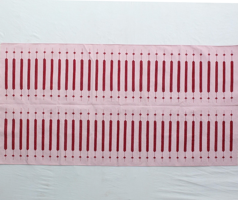 Puede incluir: Textil rectangular rosa con un patr&oacute;n repetitivo de l&iacute;neas verticales burdeos y formas de diamante. El dise&ntilde;o es sim&eacute;trico, con dos filas del patr&oacute;n. El textil es probablemente un art&iacute;culo decorativo.