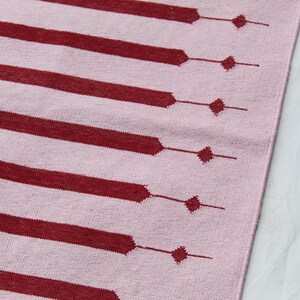 Puede incluir: Un textil rosa con un patr&oacute;n repetitivo de barras horizontales y rombos de color rojo oscuro. Las barras est&aacute;n conectadas a los rombos por l&iacute;neas finas. El textil es probablemente una alfombra o un trozo de tela.