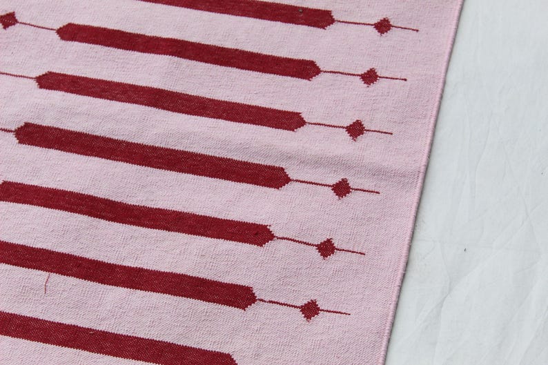 Puede incluir: Un textil rosa con un patr&oacute;n repetitivo de barras horizontales y rombos de color rojo oscuro. Las barras est&aacute;n conectadas a los rombos por l&iacute;neas finas. El textil es probablemente una alfombra o un trozo de tela.