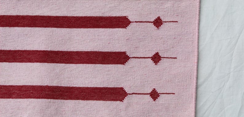 Puede incluir: Un textil tejido rosa y burdeos con un patr&oacute;n de rayas. Las rayas burdeos tienen un dise&ntilde;o geom&eacute;trico con formas de diamante. El textil es probablemente una alfombra o una tela decorativa.