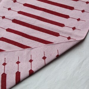 Puede incluir: Una alfombra rosa y burdeos con estampado geom&eacute;trico. La alfombra presenta rayas horizontales burdeos y l&iacute;neas verticales con forma de diamante. La alfombra est&aacute; doblada, revelando la parte inferior.