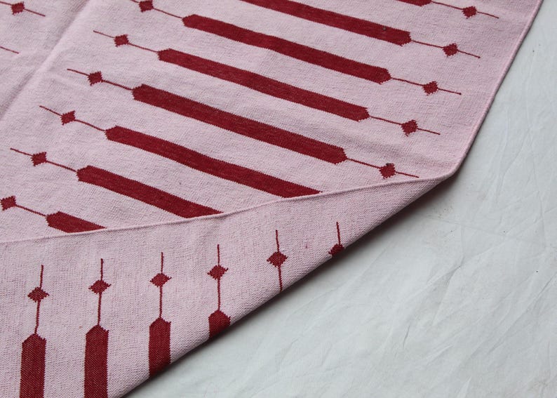 Puede incluir: Una alfombra rosa y burdeos con estampado geom&eacute;trico. La alfombra presenta rayas horizontales burdeos y l&iacute;neas verticales con forma de diamante. La alfombra est&aacute; doblada, revelando la parte inferior.