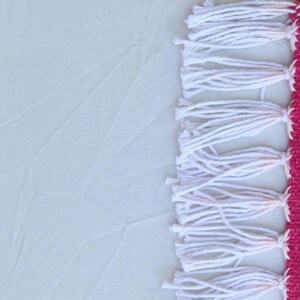 Puede incluir: Primer plano de un textil con fondo blanco y borde fucsia. El borde presenta flecos blancos. El textil es probablemente una alfombra o una pieza decorativa, con un dise&ntilde;o sencillo pero elegante.