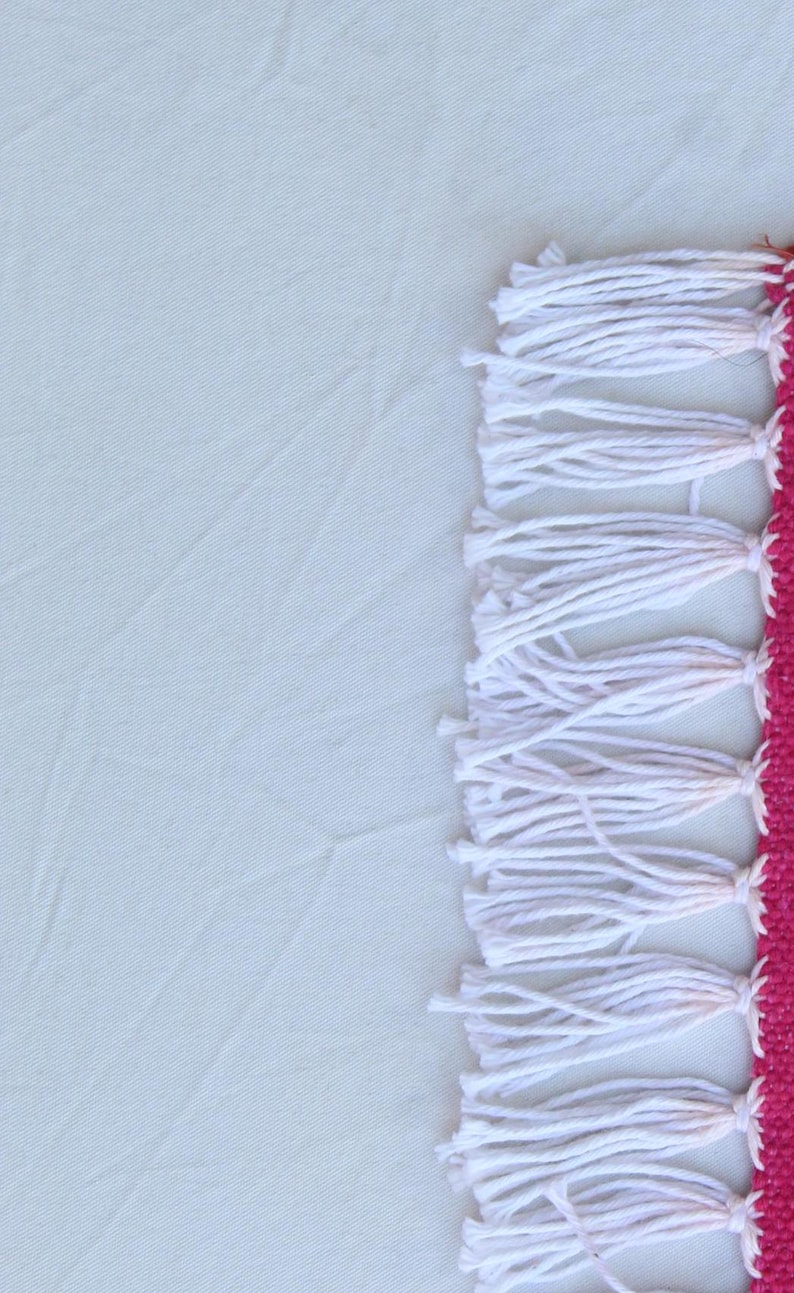 Puede incluir: Primer plano de un textil con fondo blanco y borde fucsia. El borde presenta flecos blancos. El textil es probablemente una alfombra o una pieza decorativa, con un dise&ntilde;o sencillo pero elegante.