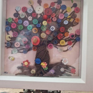 May include: A framed artwork depicting a tree made from brown yarn, decorated with a variety of colourful buttons. The background is pale pink, and the frame is white. Small decorative flowers are at the base of the tree.