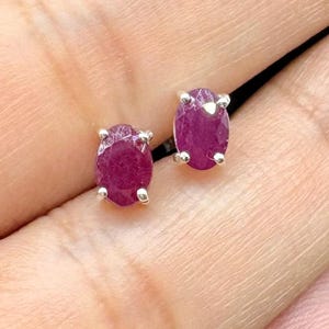 May include: A pair of oval-shaped ruby stud earrings. The rubies are a deep red color and are set in a silver metal. The earrings are displayed against a neutral background.