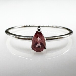 Op de afbeelding: Een zilveren ring met een peervormige, roodroze edelsteen. De ring heeft een slanke, gepolijste band en de edelsteen wordt op zijn plaats gehouden door een eenvoudige vier-klauwzetting. De ring wordt getoond op een wit oppervlak, dat licht reflecteert.