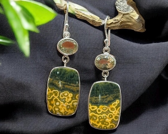 May include: A pair of dangle earrings with a silver-tone metal setting. Each earring features an oval-shaped stone at the top and a rectangular stone at the bottom. The stones are green, yellow, and brown.