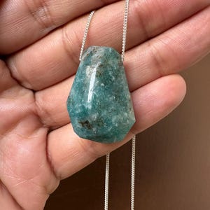 May include: A teal-colored gemstone pendant with a silver chain. The irregularly shaped stone has a polished surface with variations in color and texture. The chain is delicate and appears to be made of small, linked metal pieces. The pendant is displayed against a neutral background.