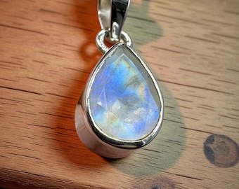 May include: A teardrop-shaped pendant with a moonstone gemstone, set in a silver metal bezel, hangs from a delicate silver chain. The moonstone displays a blue and white iridescent glow. The necklace is displayed on a wooden surface.