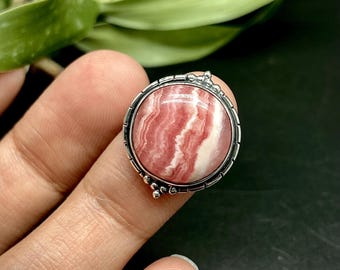May include: A round, silver ring featuring a pink and white stone with a striped pattern. The ring is held up by a finger, showcasing the intricate details of the setting and the stone's natural beauty. The ring is a piece of jewelry.