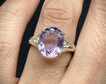 May include: A gold ring with a large, oval-cut amethyst gemstone. The amethyst is a light purple colour and is set with small accent stones on either side. The ring is shown on a person's finger.