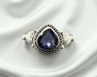 May include: A silver ring featuring a heart-shaped, deep purple gemstone. The gemstone is set within a twisted rope-like border. The ring has decorative elements on either side of the gemstone. The ring is displayed on a white, silky background.
