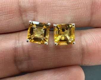 May include: A pair of square-cut, golden-yellow gemstone stud earrings. The gems are set in silver-colored metal. The earrings are displayed against a neutral background.