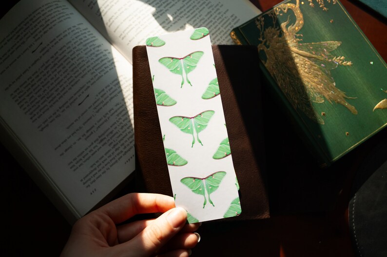 Pode incluir: Um marcador de p&aacute;gina branco com um padr&atilde;o repetido de mariposas luna verdes. O marcador de p&aacute;gina est&aacute; sendo segurado em uma m&atilde;o.