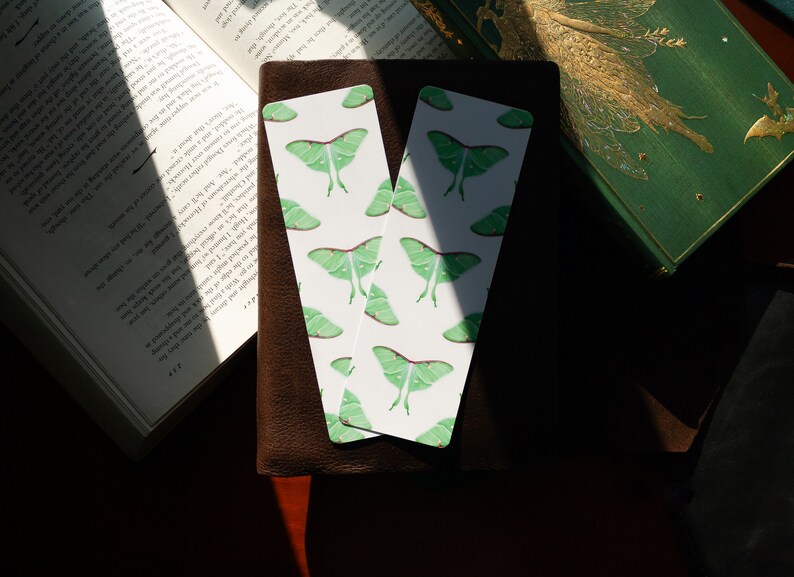 Pode incluir: Dois marcadores de p&aacute;gina brancos com um padr&atilde;o repetido de mariposas luna verdes. Os marcadores de p&aacute;gina est&atilde;o sobre uma superf&iacute;cie de couro marrom.